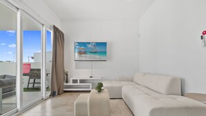 A modern living room with a large window, a flat-screen TV, a beige sofa, and a small table with a plant. - Caya Erasmus 18 (Oranjestad)