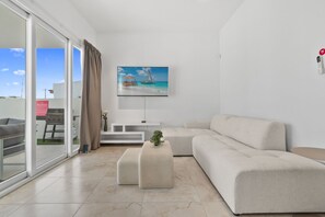 Ein modernes Wohnzimmer mit großem Fenster, einem Flachbildfernseher, einem beigen Sofa und einem kleinen Tisch mit einer Pflanze.