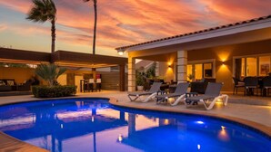 Piscina com cadeiras de descanso, um terraço coberto e um céu ao pôr do sol.