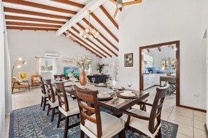 A dining area with a wooden table set for six, surrounded by chairs, with a ceiling fan above and a kitchen visible in the background.