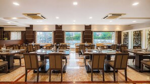 A spacious dining area with multiple tables and chairs, large windows offering a view, and a modern ceiling design. - Sumitel Central Park Hotel & Banquet (Jalpaiguri)