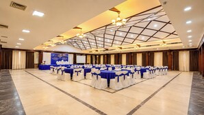 Un amplio salón de conferencias con varias mesas y sillas, un escenario con pantalla y un techo alto con tragaluces.