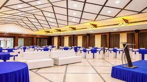 A conference room with rows of white sofas and tables, blue tablecloths, and a podium. - Sumitel Central Park Hotel & Banquet (Jalpaiguri)