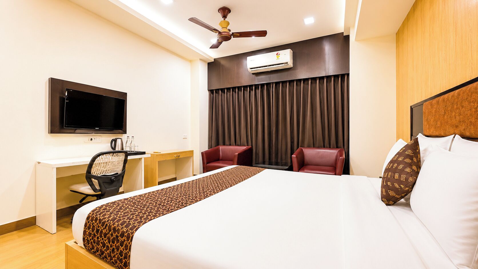 Habitación de hotel con cama, escritorio, silla, televisor, aire acondicionado y ventilador de techo.