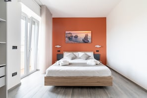 Un dormitorio moderno con una cama grande, un cuadro de un atardecer y vista al mar desde la ventana.
