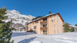 Ein mehrstöckiges Gebäude mit Balkonen, im Hintergrund eine Skipiste und verschneite Umgebung.
