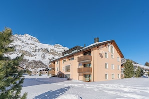 Ein mehrstöckiges Gebäude mit Balkonen, im Hintergrund eine Skipiste und verschneite Umgebung.