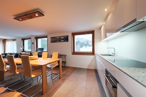 Eine moderne Küche mit Einbauherd, ein Essbereich mit Holztisch und Stühlen sowie große Fenster mit Blick ins Freie.