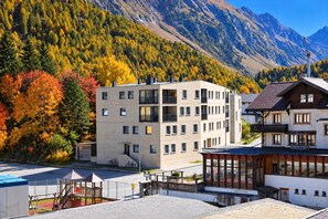 A modern building with multiple floors, balconies, and a view of a mountainous landscape with autumn foliage. - Chesa Soldanella – Stylish Vacation Rental with Valley and Mountain Views (Bregaglia)