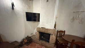 Een kamer met een stenen openhaard, een aan de muur bevestigde televisie en een eethoek met houten stoelen.