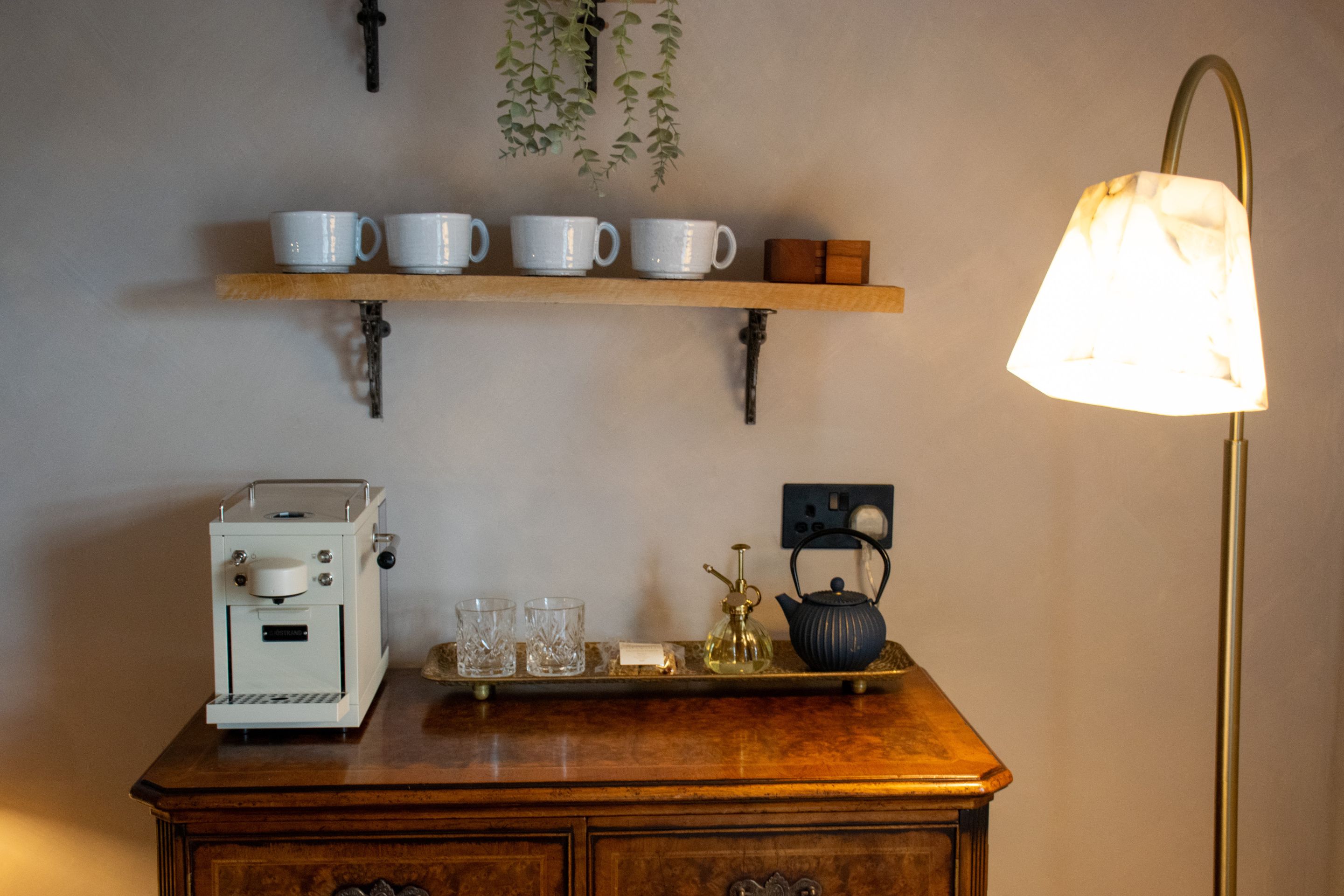 A kitchenette with a coffee maker, a tray with glasses, a teapot, and a shelf with cups.