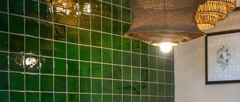 A room with green tiled walls, two mustard-colored armchairs, a small round table with a radio, and a hanging woven light fixture.