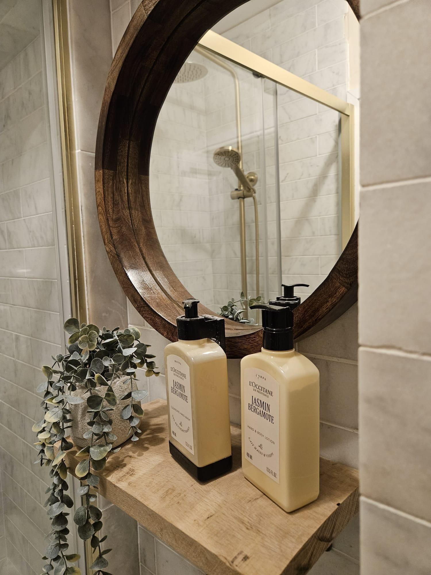 A bathroom with a wooden shelf holding two bottles of body lotion and a mirror with a wooden frame.