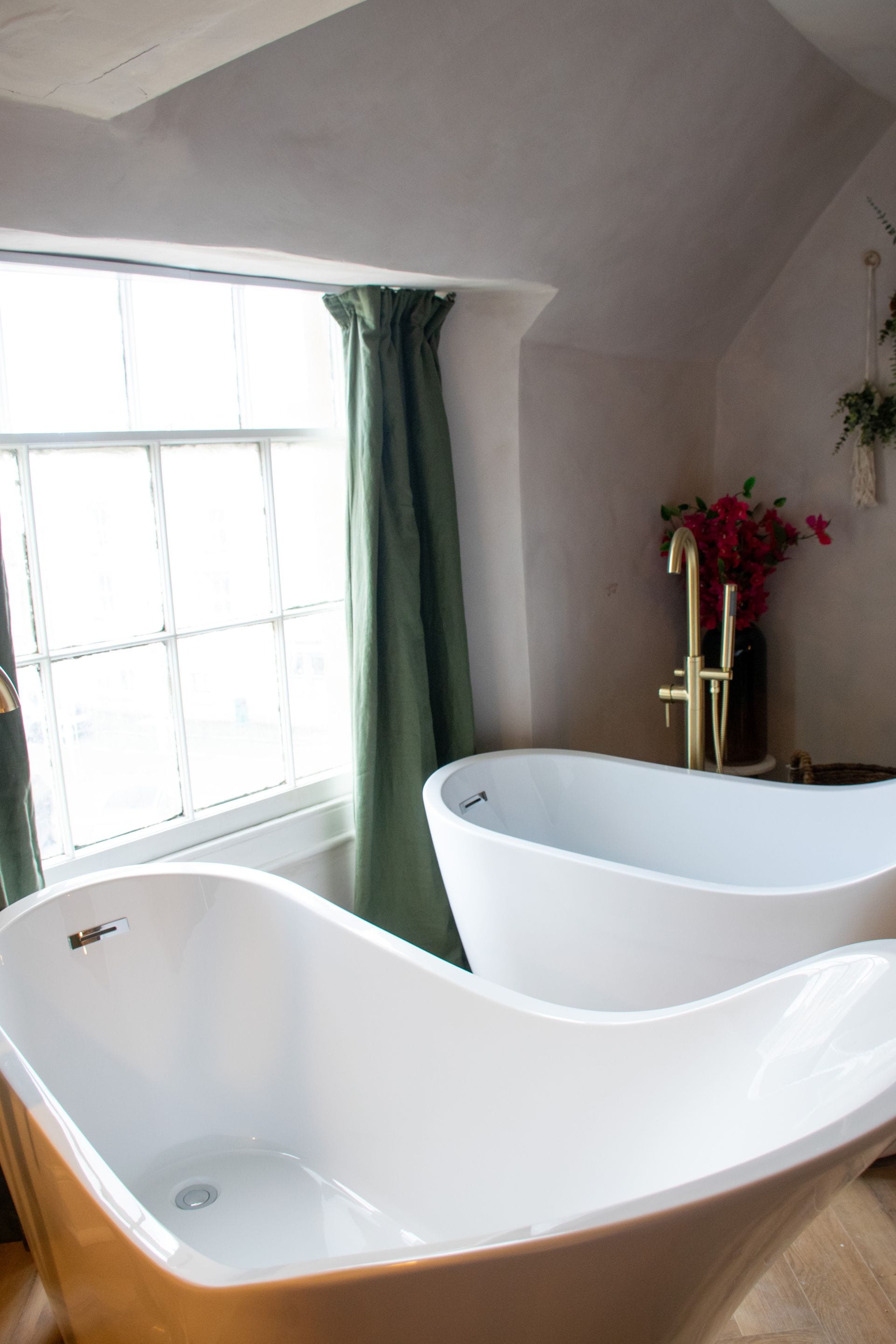 A bathroom with two freestanding tubs, a window with curtains, and a vase with flowers.