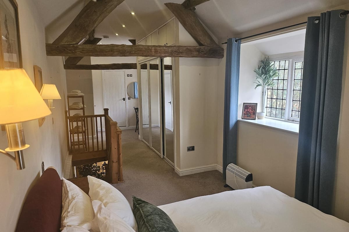 A bedroom with a bed, a chair, a window with curtains, a radiator, and a hallway leading to a door.