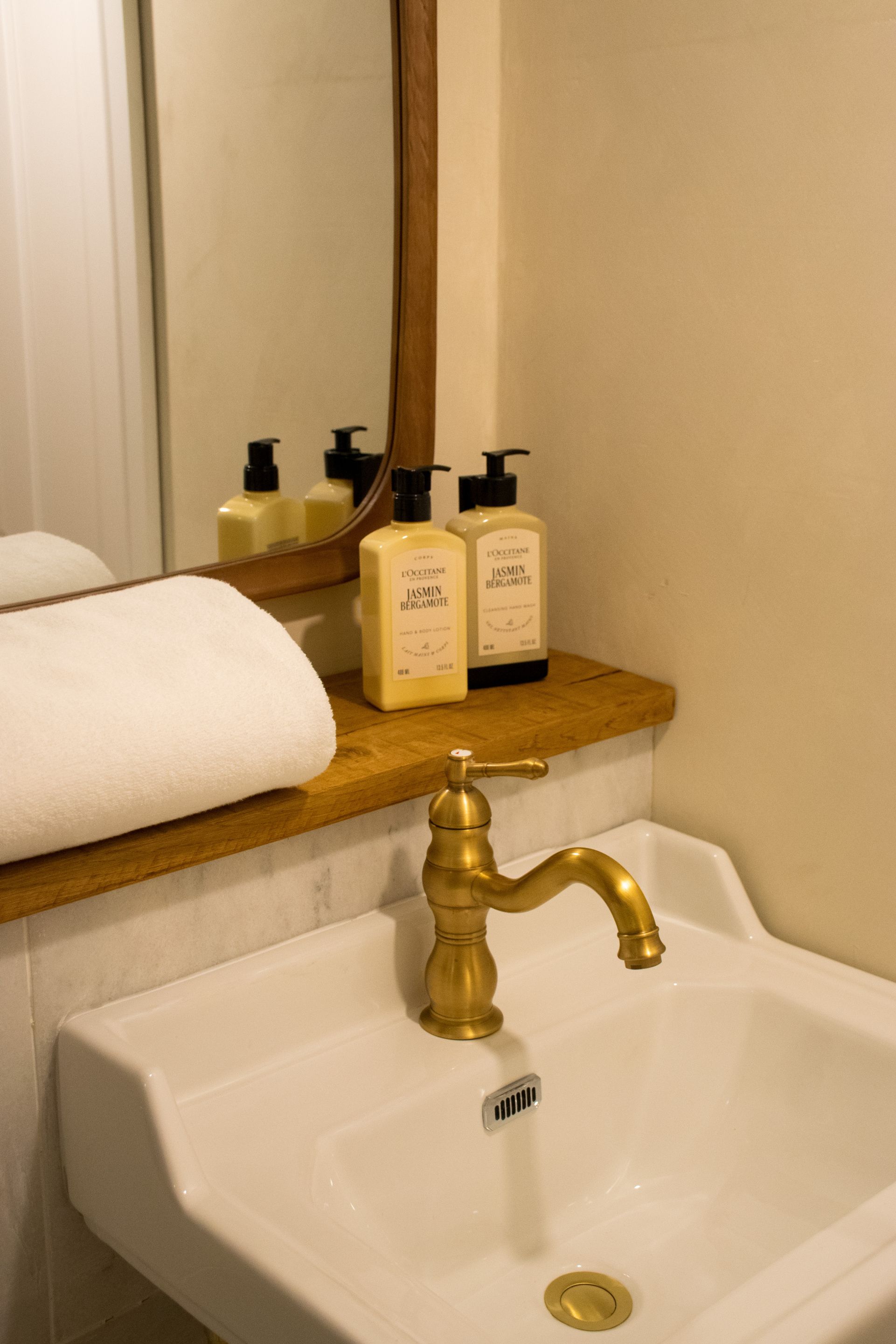 Ein Badezimmer mit einem weißen Waschbecken, einem goldfarbenen Wasserhahn und zwei Flaschen L'OCCITANE Körperlotion auf einem Holzregal.
