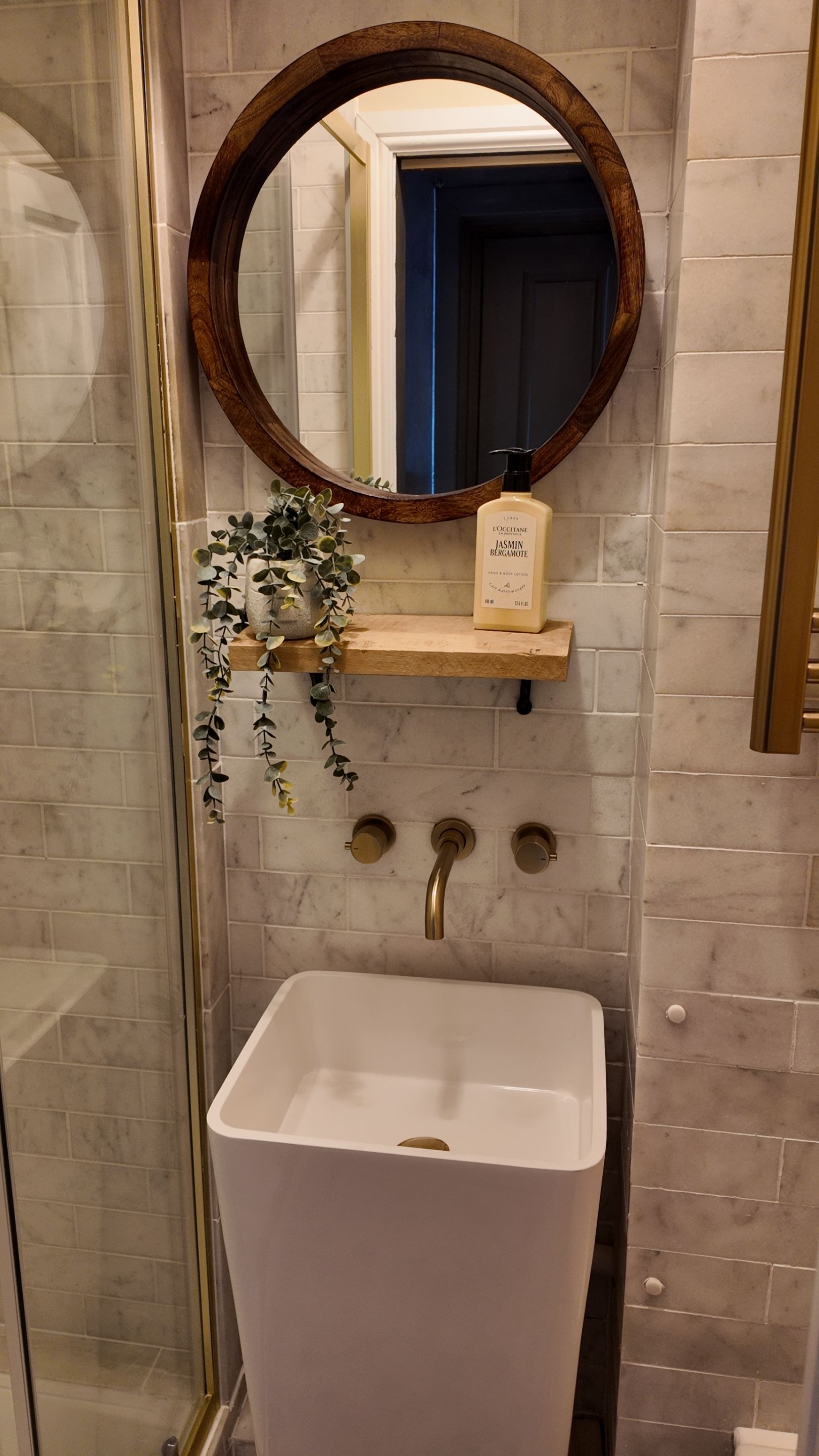 Un baño con un lavamanos blanco, un estante de madera con una botella y un espejo con marco de madera.