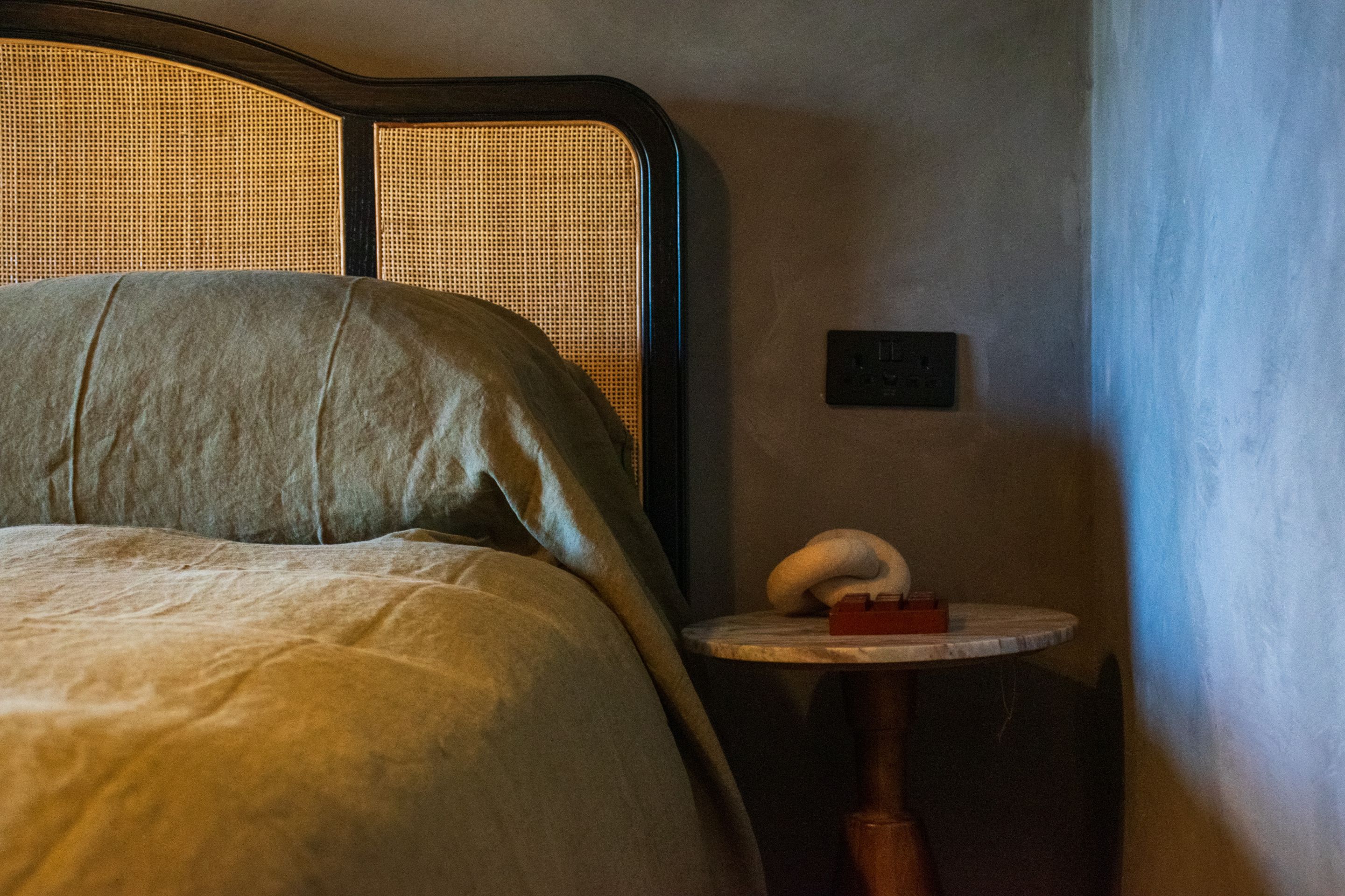 A bed with a headboard, a bedside table with a lamp, and a wall socket.