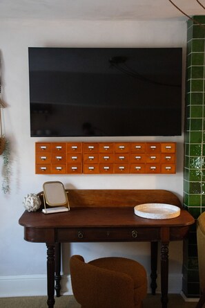 A room with a large flat-screen TV mounted on the wall, a wooden sideboard with multiple drawers, a small round table with a plate, and a brown chair. - Cotswold Townhouse - Adults Only (Stow-on-the-Wold)