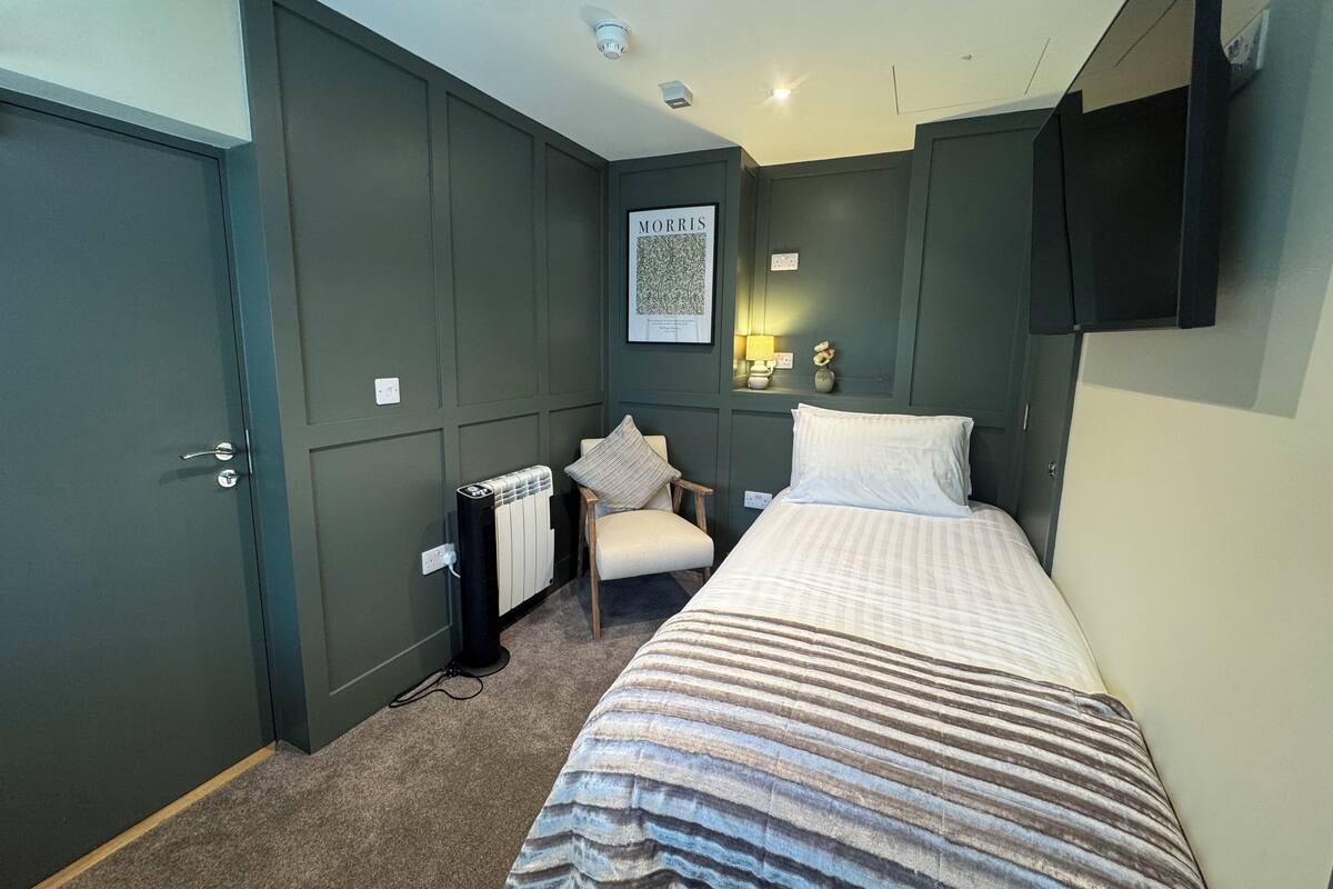 Une chambre à coucher comprenant un lit, une chaise, un radiateur, un téléviseur fixé au mur et un tableau encadré.