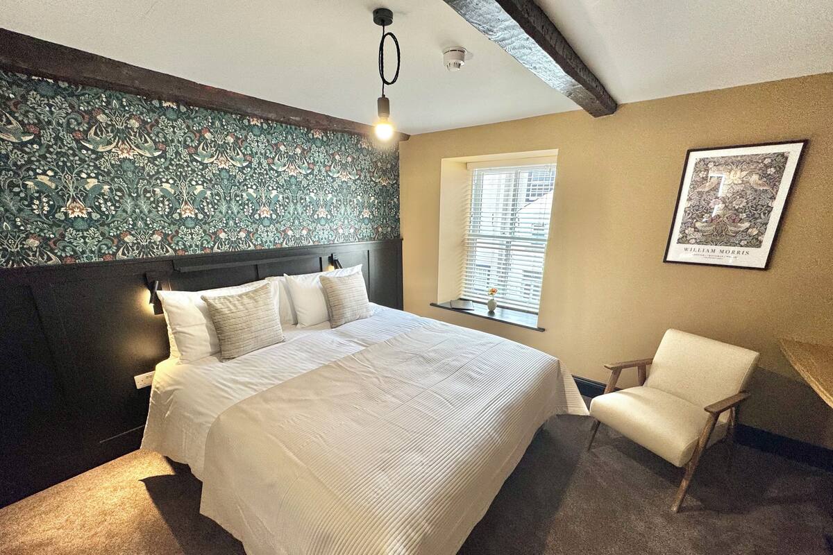 Une chambre avec un grand lit, une chaise, une fenêtre avec des stores, un luminaire fixé au mur et un tableau encadré.