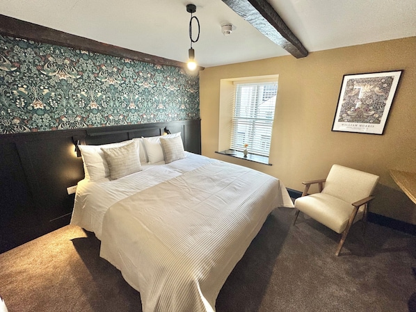 A bedroom with a large bed, a chair, a window with blinds, a wall-mounted light, and a framed picture.