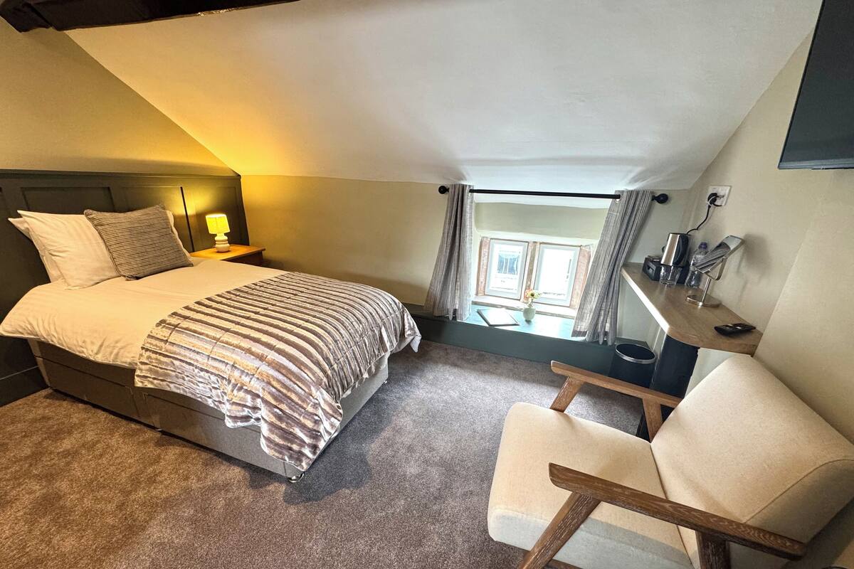 Une chambre Ă coucher avec un lit, une table de chevet, une lampe, une chaise et une fenĂŞtre avec des rideaux.