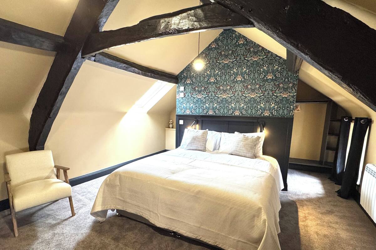Une chambre avec un plafond en pente, un lit doté d’une tête de lit à motifs, une chaise et un radiateur.