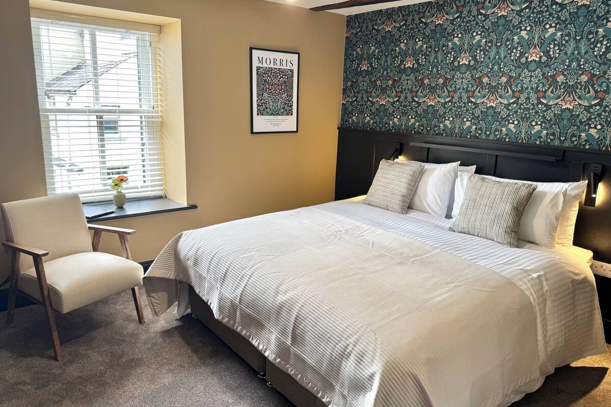 Une chambre à coucher comprenant un lit, une chaise, une fenêtre avec des stores, un tableau encadré au mur et un papier peint à motifs.