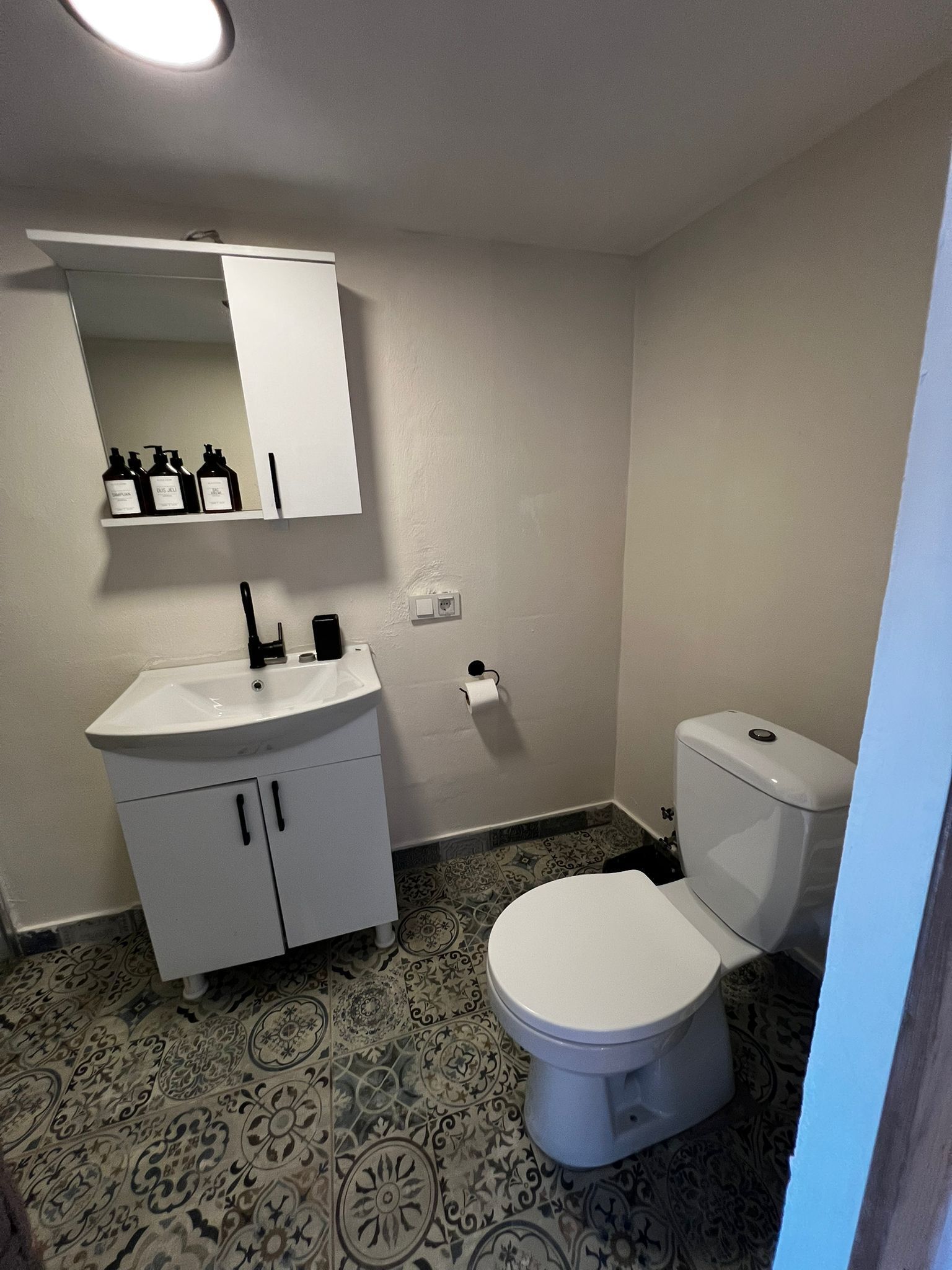 Un baño con lavamanos blanco, espejo, inodoro, baldosas de suelo estampadas y un armario blanco.