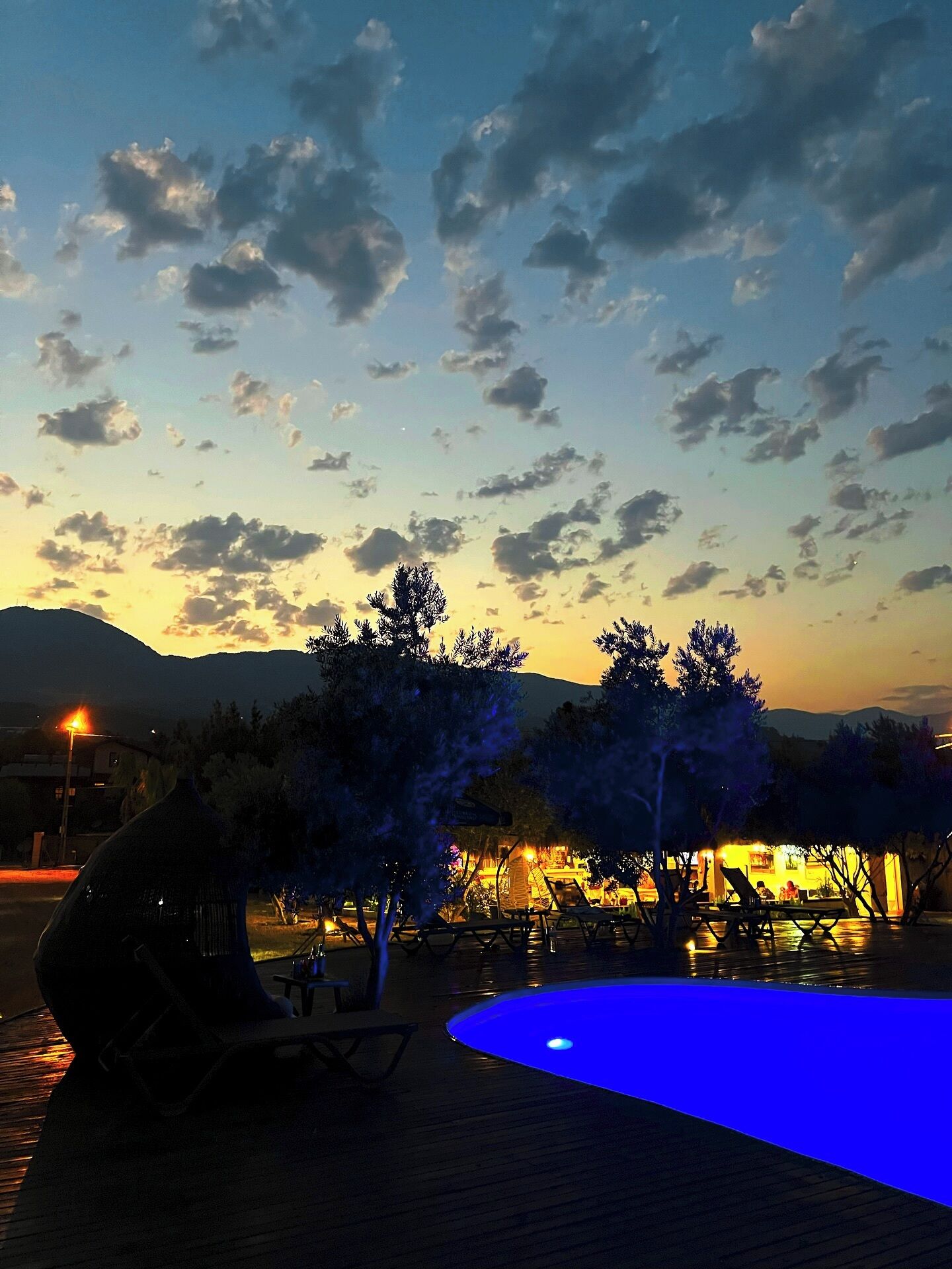 Un espace piscine avec un bord illuminé de lumière bleue, des arbres et un bâtiment éloigné dont les lumières sont visibles.