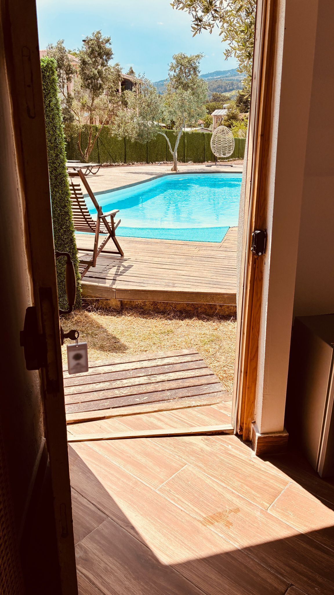 Puerta abierta que da a un patio trasero con piscina, terraza de madera y sillones de descanso.