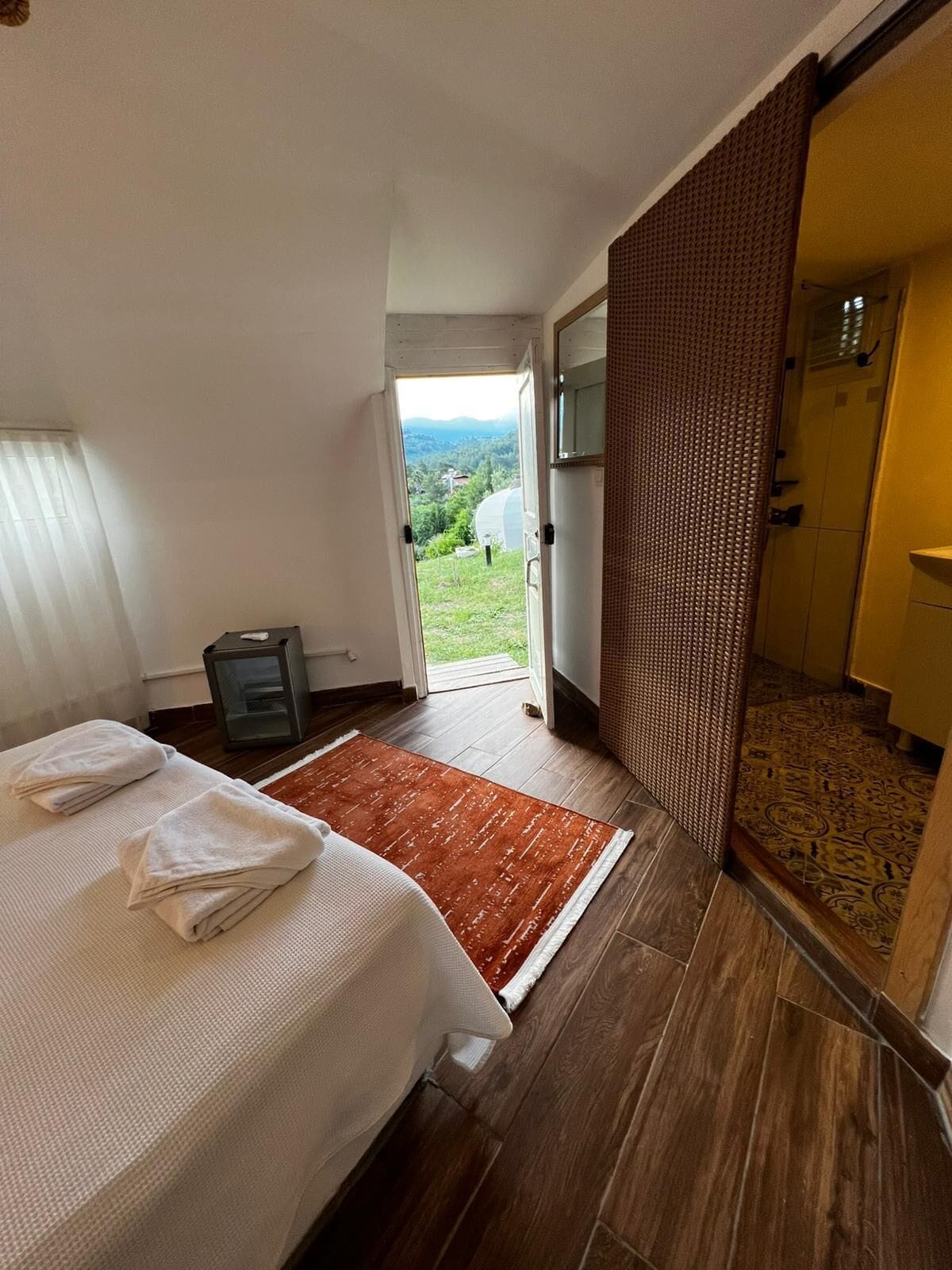 A bedroom with a bed, a small table, a rug, a shower, and a view of the outdoors.