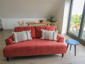 A red sofa with two pillows, a small round table, a wooden dining table, and a view of greenery outside. - Glynllifon 8 (Marianglas)