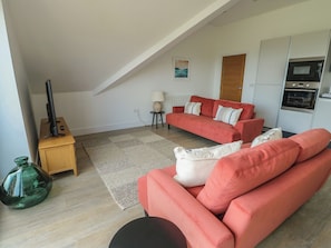 A living room with a red sofa, a wooden cabinet, a green vase, and a kitchen area with a microwave. - Glynllifon 8 (Marianglas)