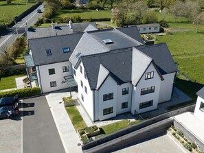 Ein großes, weißes Gebäude mit vielen Fenstern und einem geneigten Dach, umgeben von einer gepflasterten Auffahrt und einer Rasenfläche.