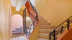 Un escalier avec une rampe en métal, une porte sur la gauche et, en haut, une vue sur une autre pièce.
