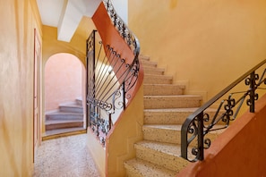Una escalera con barandilla metálica, una puerta a la izquierda y vista de otra habitación al final.