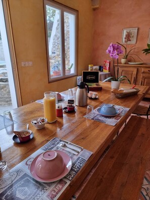 Una mesa de madera con un desayuno dispuesto, que incluye una tetera rosa, un vaso de zumo de naranja y un hervidor.