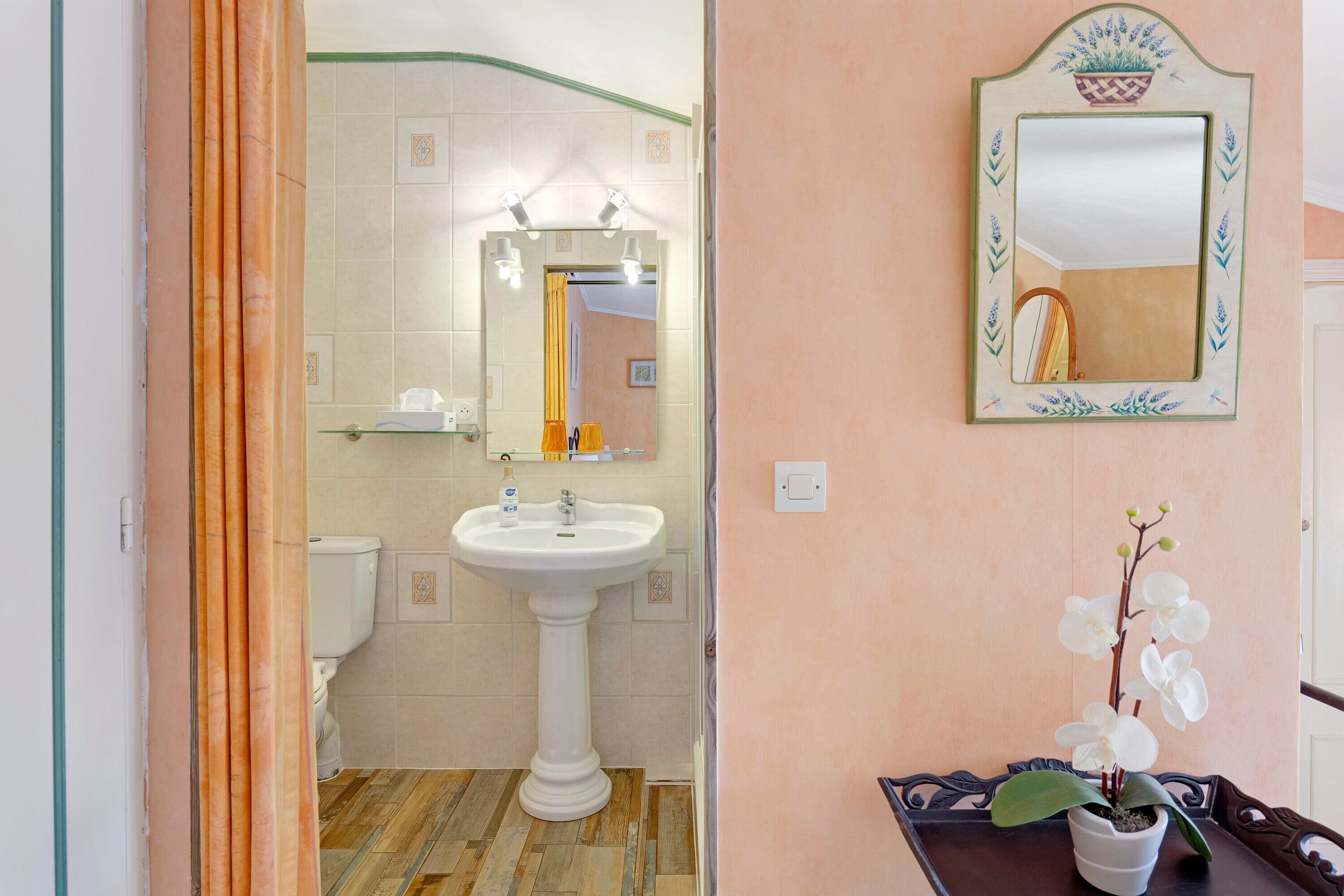A bathroom with a pedestal sink, a mirror with a decorative frame, and a potted orchid on a table.