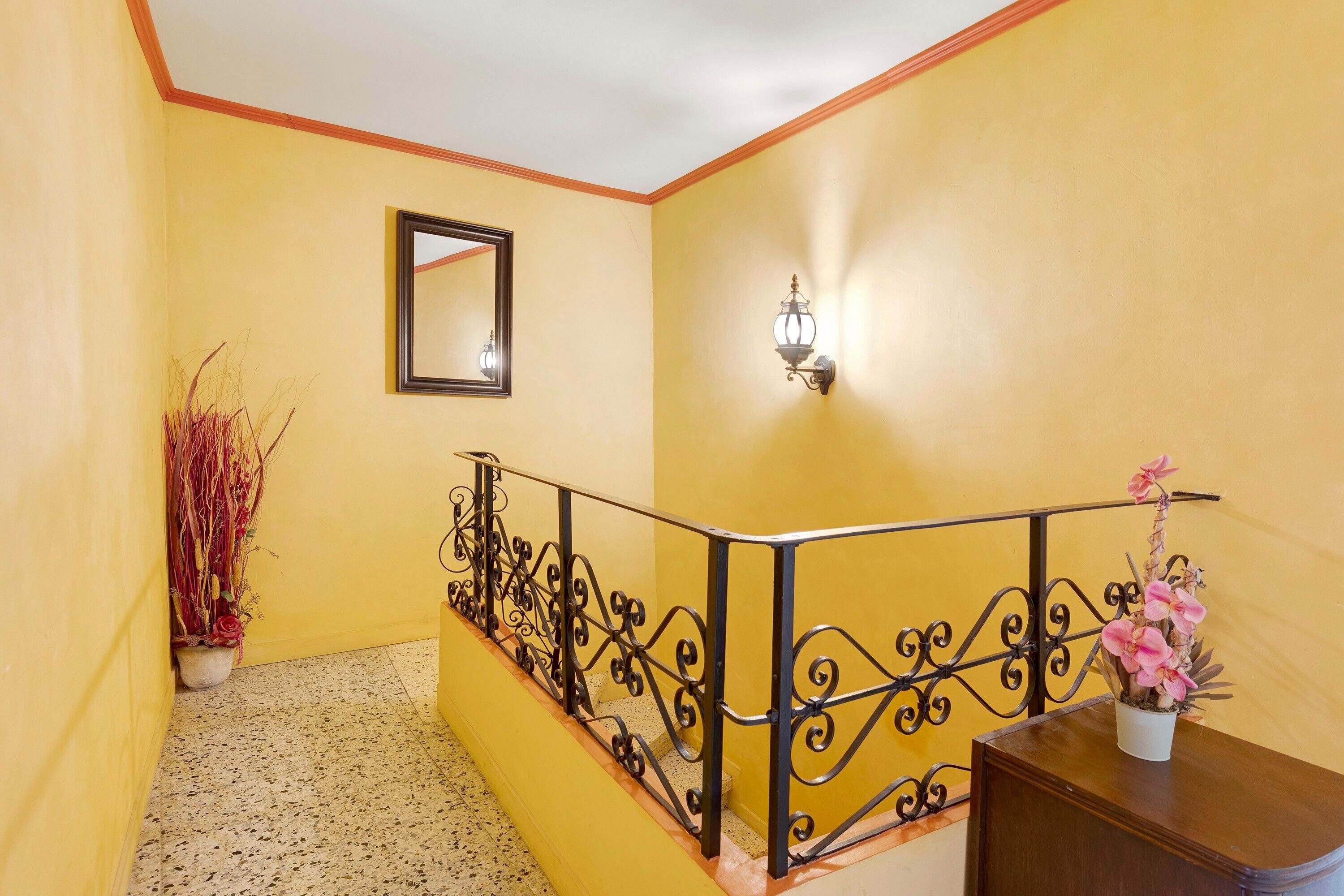 A hallway with yellow walls, a metal railing, a potted plant, a mirror, and a wall-mounted light fixture.