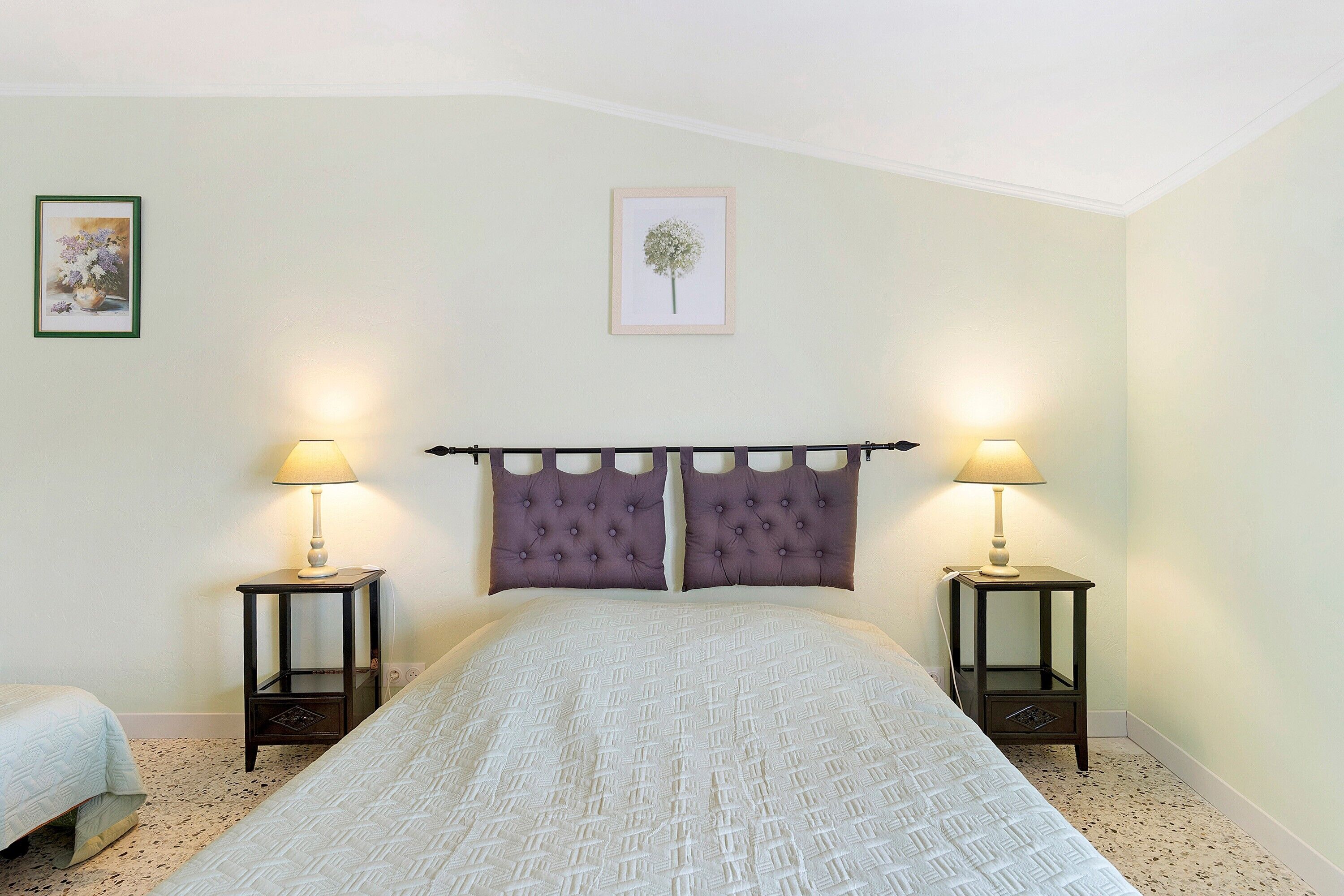 A bedroom with a bed, two bedside tables with lamps, a headboard, and two framed pictures on the wall.