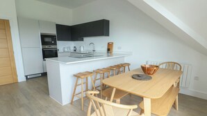 A modern kitchen with a central island, bar stools, and a dining table with chairs. - Glynllifon 9 (Marianglas)