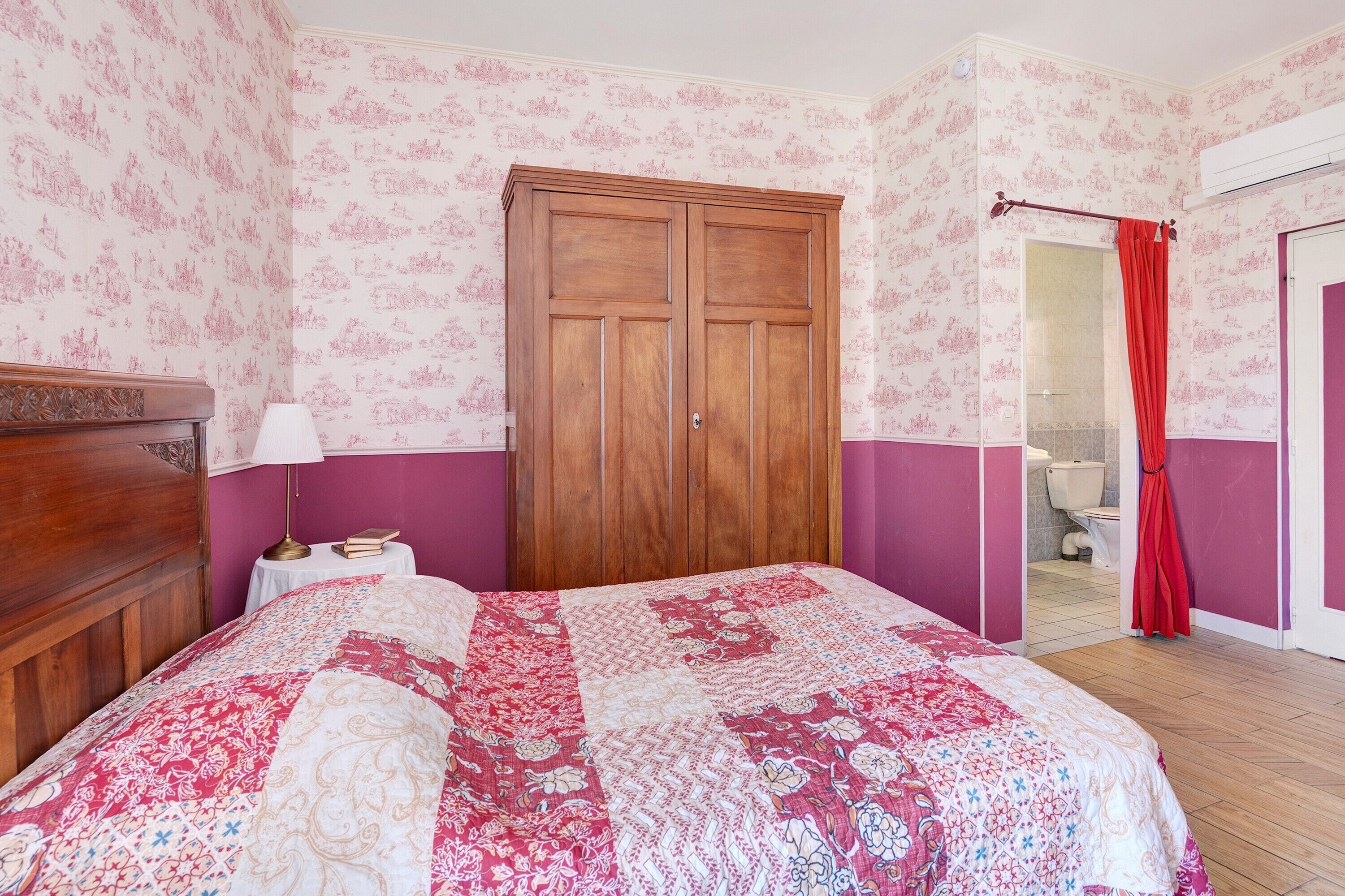 Une chambre à coucher avec une armoire en bois, un lit recouvert d’une courtepointe à motifs floraux, une table de chevet avec une lampe, et une salle de bain visible à travers une porte ouverte.