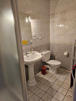 Un baño con lavamanos pedestal, inodoro, repisa de vidrio y bote de basura rojo.