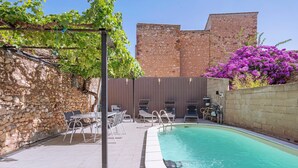 A swimming pool with a stone wall, a pergola with vines, and a brick building in the background.