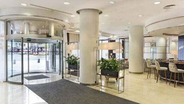 A modern hotel lobby with a revolving door, a reception desk, and a seating area with chairs. - Hotel Ini (Madrid)