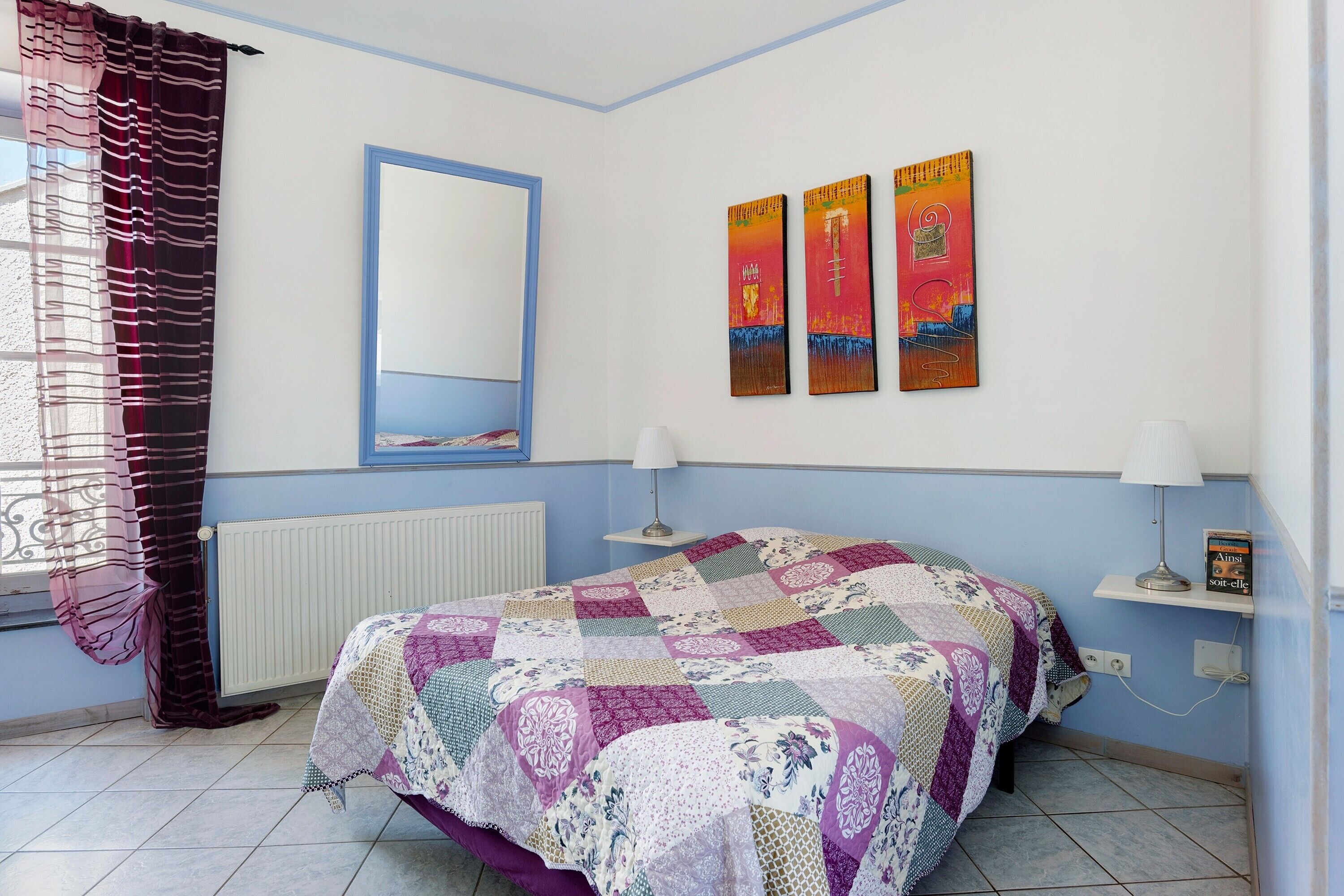 Een slaapkamer met een bed, een raam met gordijnen, een radiator, een spiegel aan de muur en drie schilderijen.