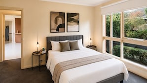A bedroom with a large bed, two bedside tables, a headboard, and a window with a view of greenery. - Family-Friendly 2BR Villa with Private Courtyard (Malvern East)