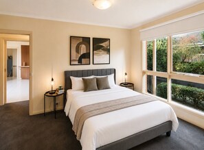 Een slaapkamer met een groot bed, twee nachtkastjes, een hoofdeinde en een raam met uitzicht op groen.